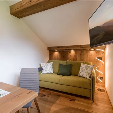 Ein gemütlicher Wohnraum mit einem grünen Sofa und einem Holztisch. An der Wand hängt ein Fernseher, und sanfte Beleuchtung sorgt für eine einladende Atmosphäre.