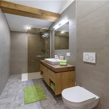 Ein modernes Badezimmer mit einem Holzregal und einer großen Dusche. Es enthält eine Waschbeckenmögel und grüne Handtücher für einen frischen Akzent.