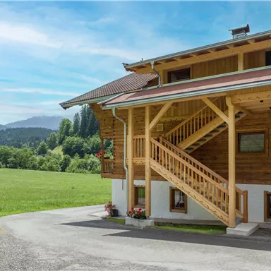 Ein gemütliches Holzhaus mit einer Außentreppe und blühenden Pflanzen. Im Hintergrund erstrecken sich grüne Wiesen und Berge unter einem klaren Himmel.