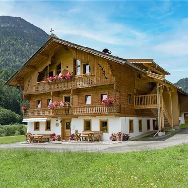 Ein traditionelles Chalet aus Holz mit Balkonen und Blumenkästen. Die Umgebung ist grün und von Bergen umgeben.