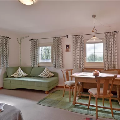 Ein gemütliches Wohnzimmer mit einem grünen Sofa und einem Esstisch. Durch die Fenster fällt viel Tageslicht und die Vorhänge verleihen dem Raum eine einladende Atmosphäre.