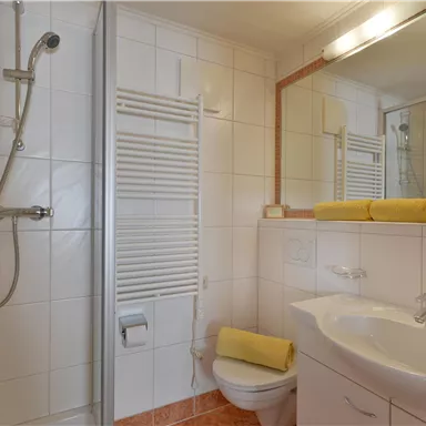 Ein modernes Badezimmer mit Dusche und weißen Fliesen. Auf dem Waschbecken liegt ein gelbes Handtuch.