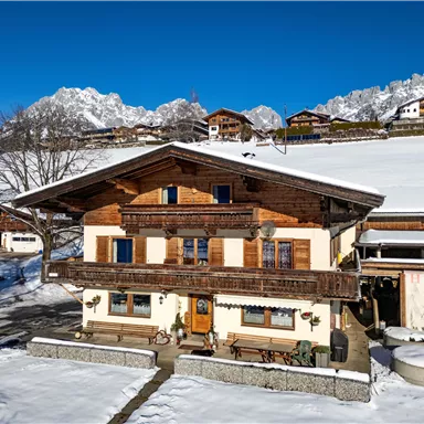 A traditional wooden house in the snow, surrounded by mountains. The sky is clear and blue, which enhances the wintry atmosphere.