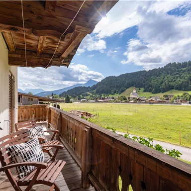 A balcony with two wooden chairs and a view of green meadows and mountains. The sky is clear with some clouds.
