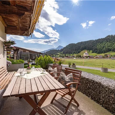 A cozy balcony with a wooden table and chairs. In the background, green meadows and mountains can be seen under a clear sky.