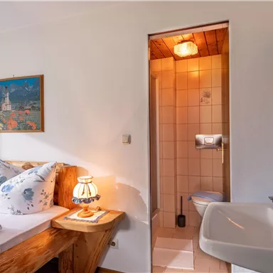 A cozy bedroom with two beds, pretty pillows, and a small table. In the background, a bathroom with a shower and sink can be seen.