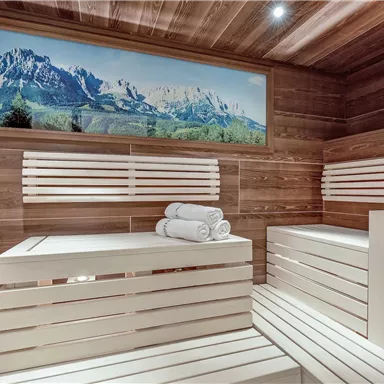 A modern sauna area with wooden paneling and white benches. In the background, a beautiful mountain panorama can be seen.