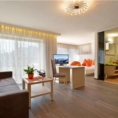 Ein modernes Wohnzimmer mit gemütlichem Sofa, einem Tisch und großen Fenstern. Die orangefarbenen Kissen und Pflanzen verleihen dem Raum eine einladende Atmosphäre.