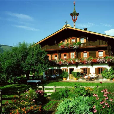 A traditional Bavarian house with a beautiful balcony full of flowers. The surroundings are green and idyllic, surrounded by trees and blooming garden plants.