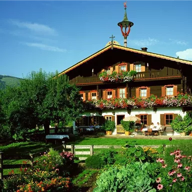Ein traditionelles, bayerisches Haus mit einem schönen Balkon voller Blumen. Die Umgebung ist grün und idyllisch, umgeben von Bäumen und blühenden Gartenpflanzen.