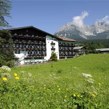Ein gemütliches Hotel mit Balkonen, umgeben von einer grünen Wiese und Bergen im Hintergrund. Der Himmel ist klar und die Natur ist einladend.