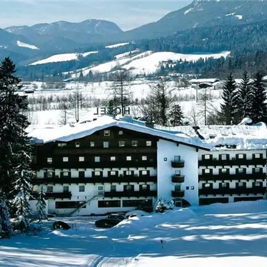 An elegant hotel in the snow with a picturesque mountain panorama in the background. Surrounded by tall, snow-covered fir trees, the winter landscape stretches out.