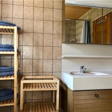 Ein modernes Badezimmer mit braunen Fliesen und einem großen Spiegel. Handtücher sind ordentlich auf einem Holzregal gestapelt.