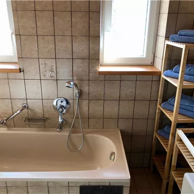 Ein sauberes Badezimmer mit einer Badewanne und hellen Fliesen. Auf einem Holzregal liegen saubere Handtücher.