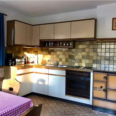 Eine gemütliche Küche mit Holzfurnier-Elementen und einer Fliesenwand in Erdtönen. Am Tisch liegt ein kariertes Tuch und die Küche ist gut beleuchtet.