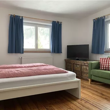 Ein helles Schlafzimmer mit einem großen Bett und karierten Bettwaren. Neben dem Bett steht ein gemütlicher Sessel und ein Fernseher an der Wand.