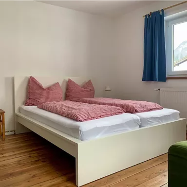 Ein gemütliches Schlafzimmer mit einem weißen Bett und karierten Kissen. Das Zimmer hat Holzfußboden und ein Fenster mit blauen Vorhängen.
