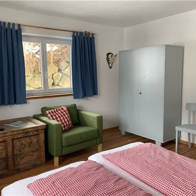 Ein gemütliches Zimmer mit einem Bett, einem grünen Sessel und einem Holztisch. Die Fenster sind mit blauen Vorhängen dekoriert.