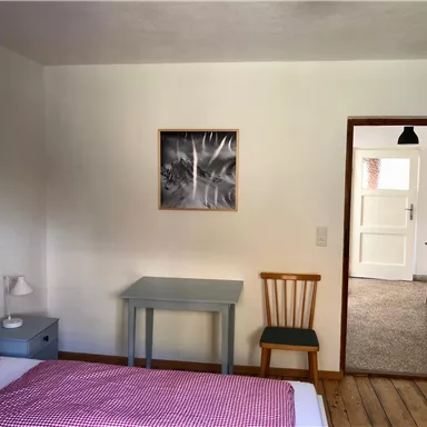 Ein einfaches Schlafzimmer mit einem Bett, einem kleinen Tisch und einem Stuhl. Im Hintergrund ist ein weiteres Zimmer mit einer offenen Tür sichtbar.
