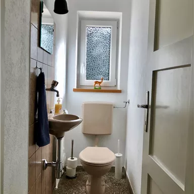 Ein kleines, helles Badezimmer mit einem Fenster und einem Waschbecken. Die Wände sind hell gestrichen, und es gibt einige Sanitäranlagen.