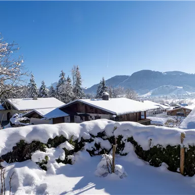 Eine winterliche Landschaft mit schneebedeckten Dächern und Bäumen. Im Hintergrund sind sanfte Berge und ein klarer blauer Himmel zu sehen.