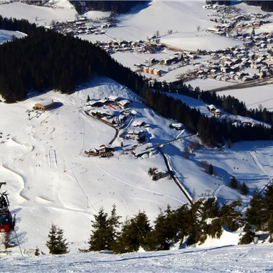 Eine schneebedeckte Landschaft mit einer Seilbahn und einem kleinen Skigebiet. Im Hintergrund sind Häuser und Wälder sichtbar.