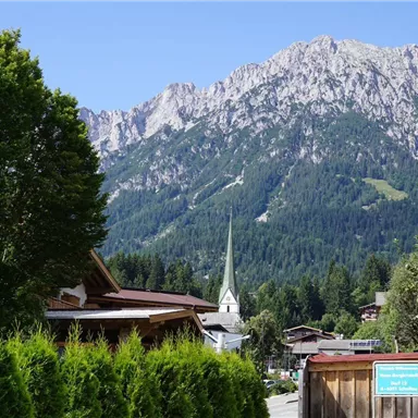A picturesque mountain landscape with high peaks and lush green forest. In the foreground, wooden houses and a church with a pointed tower are visible.