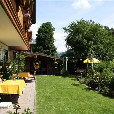 Ein schöner Garten mit einem gepflegten Rasen und vielen Pflanzen. Neben dem Haus stehen Tische mit gelben Tischdecken und eine Sonnenschirm.