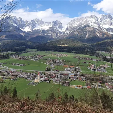 A picturesque landscape with a small village nestled among green meadows and snow-covered mountains. The sky is partly cloudy, which makes the scene especially impressive.