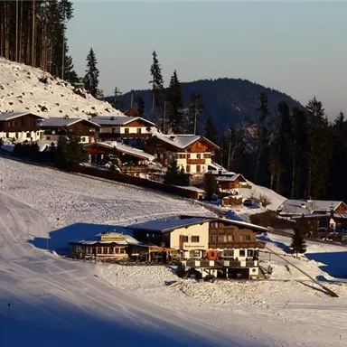 Eine verschneite Landschaft mit gemütlichen Hütten, die auf einem Hang stehen. Die Umgebung ist von Bäumen und Bergen umgeben und strahlt eine idyllische Winteratmosphäre aus.