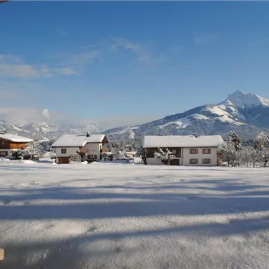 A winter landscape with snow-covered huts and mountains in the background. The sky is clear and blue.