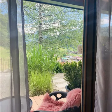 A beautiful view from a window onto a green landscape with plants. On the terrace is a comfortable chair with a pink cushion.