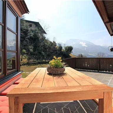 A cozy terrace with a wooden table and chairs. In the background, mountains and a sunny landscape with a green garden can be seen.