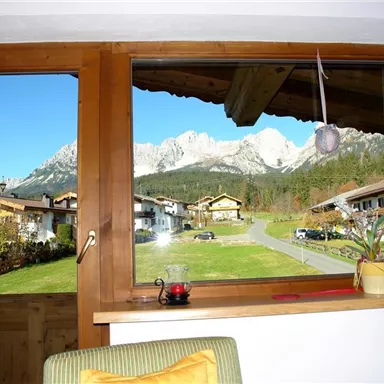 View from a window of a picturesque mountain landscape with clear blue sky. In the foreground, traditional houses and a green meadow can be seen.