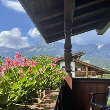 Ein malerischer Balkon mit bunten Blumen und einem Blick auf majestätische Berge. Der Himmel ist klar und die Landschaft ist grün und einladend.