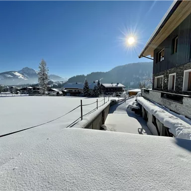 Eine winterliche Landschaft mit schneebedeckten Feldern und Bergen im Hintergrund. Die Sonne scheint hell am blauen Himmel und beleuchtet die Umgebung.