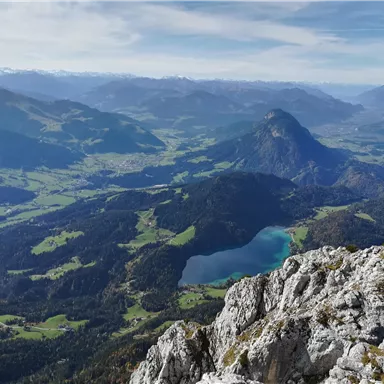 Eine malerische Landschaft mit Bergen und grünen Wiesen. Im Vordergrund sind Felsen, während im Hintergrund ein klarer See und weitere Berge zu sehen sind.