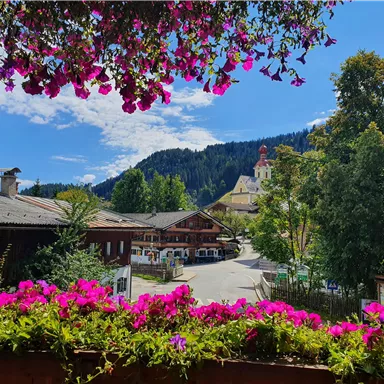 A picturesque landscape with colorful flowers in the foreground. In the background, wooden buildings and a church tower can be seen against a mountain backdrop.
