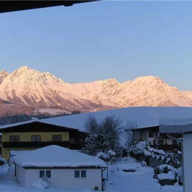 Eine winterliche Berglandschaft mit schneebedeckten Gipfeln und einem klaren Himmel. Im Vordergrund sind schneebedeckte Häuser und Bäume zu sehen.