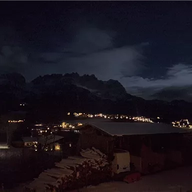 A nighttime mountain landscape with a clear moon. The lights of the village shimmer gently in the dark.