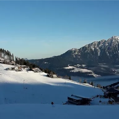 Eine verschneite Landschaft mit Bergen im Hintergrund. Kleine Hütten sind entlang des Hanges verteilt.
