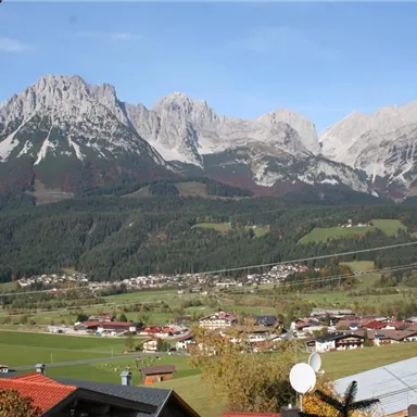 A picturesque mountain landscape with high mountains and a clear blue sky. In the foreground, there are small houses and a green valley.