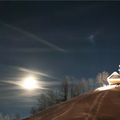 A snowy landscape at night with a bright moon. In the foreground, there is a small building on a hill, surrounded by trees.