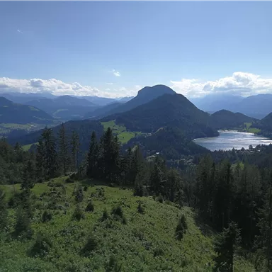 A panoramic view of green mountains and a serene lake. In the background, more mountain peaks and a blue sky can be seen.