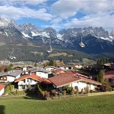 Eine malerische Landschaft mit idyllischen Häusern und majestätischen Bergen im Hintergrund. Seilbahnen fahren über die Szene und der Himmel ist leicht bewölkt.
