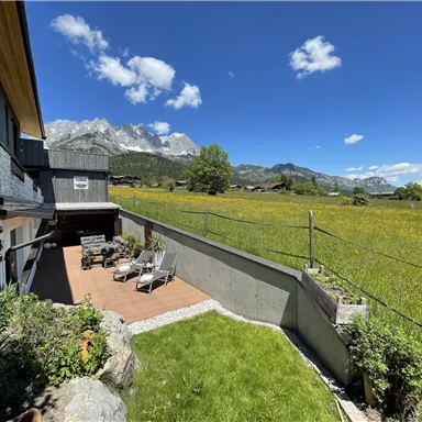 Ein schöner Garten mit Liegestühlen und einer Aussicht auf die umliegenden Berge. Der Himmel ist klar und die Wiese ist grün und blühend.