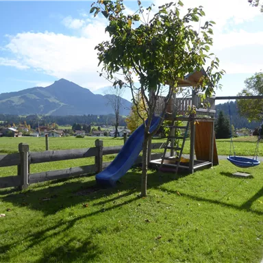 A playground with a slide and a swing in a green meadow. In the background, mountains and a blue sky can be seen.