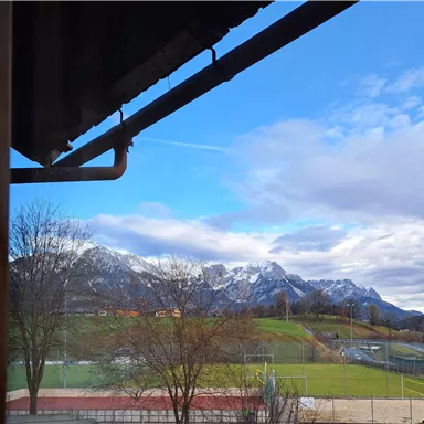 Eine Berglandschaft mit schneebedeckten Gipfeln unter einem blauen Himmel. Im Vordergrund sind Bäume und Sportplätze zu sehen.
