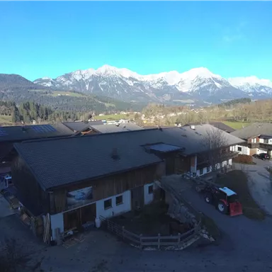 A rural scene with buildings and a clear blue sky. In the background, majestic mountains can be seen.