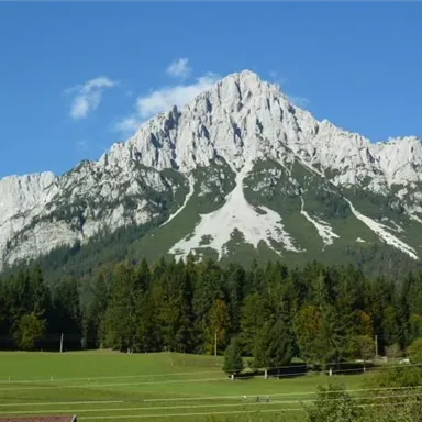 Ein majestätischer Berg mit einer beeindruckenden Felsenstruktur. Im Vordergrund erstrecken sich grüne Wiesen und Wälder.